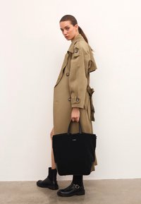 Femeie îmbrăcată într-o trench coat bej și cizme negre, ținând o geantă mare neagră, stând în fața unui perete alb simplu.