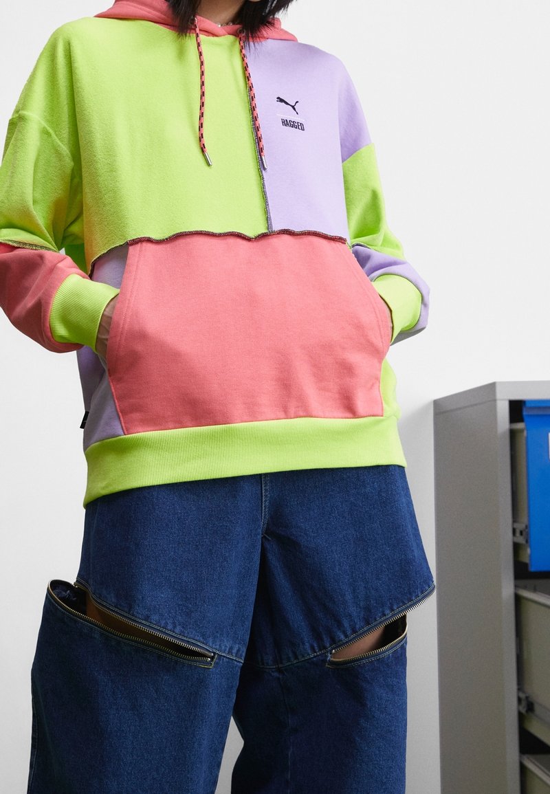 Colorful hooded sweatshirt in green, purple, and pink, featuring a cropped design, embroidered logo, and adjustable drawstrings. Worn with dark denim pants with zippers.