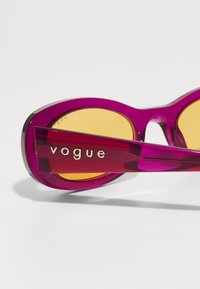 Ochelari de soare mov cu lentile galbene și o finisare lucioasă. Brațul are inscripționată cu litere aurii cuvântul "vogue". Accentuati cu un detaliu texturat.