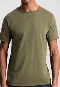 Tricou scurt de culoare verde măslin, dintr-un material moale și texturat, cu decolteu rotund și croială lejeră, având un tiv ușor curbat.