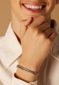 Zilveren chain armband met in elkaar grijpende schakels, gedragen op een hand tegen een lichte stoffen achtergrond. Gladde textuur en gepolijst oppervlak.