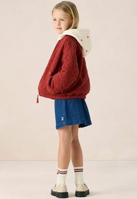 Rode gewatteerde jas met een witte capuchon met rode stippen, gecombineerd met een denim rok en crèmekleurige plateau-schoenen, aangevuld met gestreepte sokken.