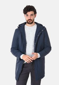 Parka impermeabile di colore blu navy con cappuccio, caratterizzata da una chiusura frontale con zip e bottoni a pressione. Indossata sopra un leggero maglione di maglia e pantaloni scuri.