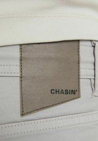 Étiquette en cuir marron avec le texte "CHASIN'" cousue sur un pantalon beige clair, partiellement recouverte par une ceinture en tissu crème.