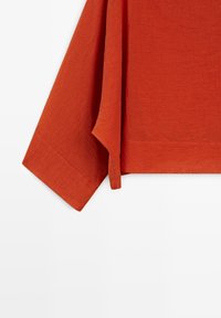 Langarmig shirt in roestkleur met zichtbare stikseldetails op de manchetten en gestructureerde stof tegen een witte achtergrond.