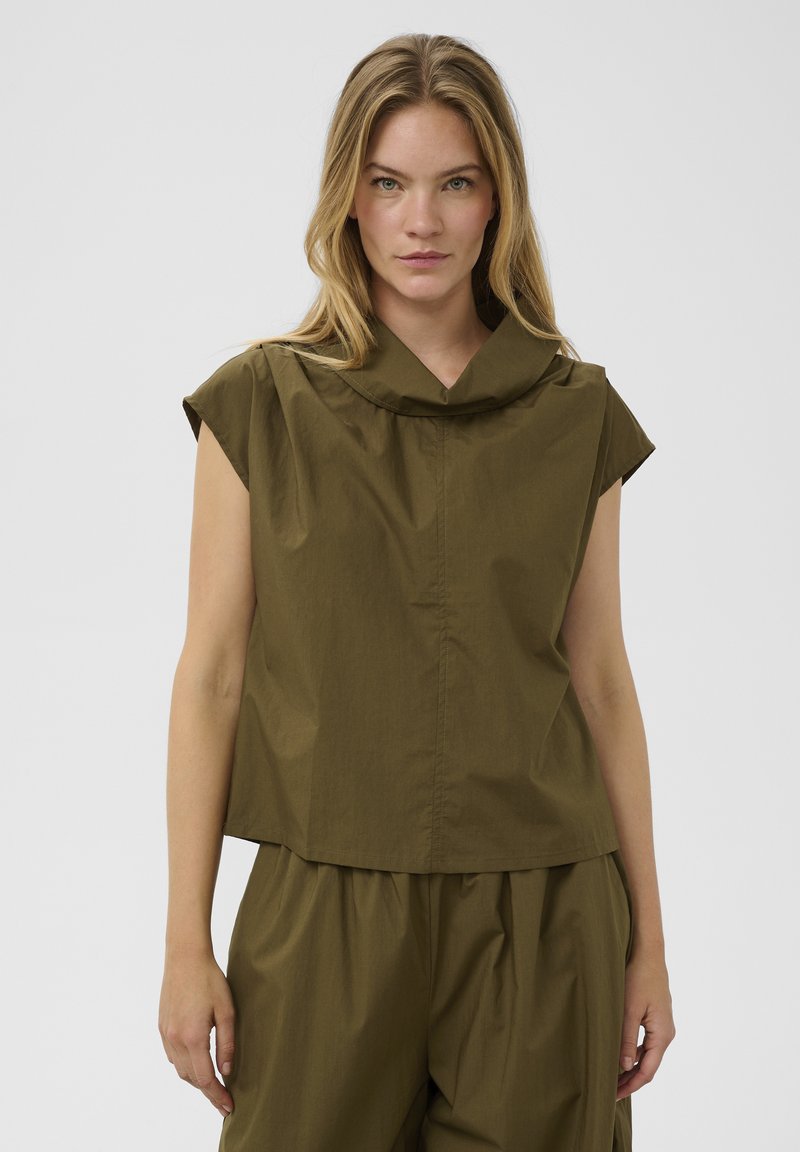 Femme aux cheveux blonds portant un haut sans manches vert olive et un pantalon ample assorti, debout devant un fond uni.