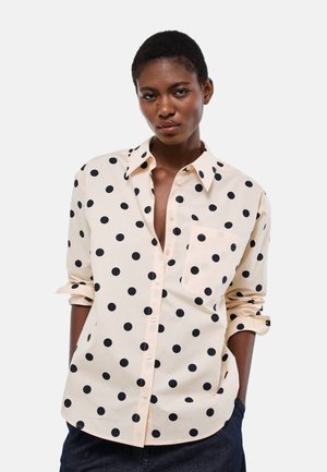 Femme portant une chemise beige clair à boutons avec de grands pois noirs, manches retroussées, et pantalon foncé sur un fond uni.