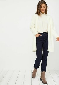 Długi biały kardigan z miękkiego materiału, zestawiony z jasną bluzką i ciemnoniebieskimi dżinsami, uzupełniony beżowymi botkami za kostkę.