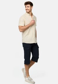 Chemise à boutons à manches courtes beige clair, shorts cargo bleu marine foncé et baskets grises. Tenue décontractée avec poches sur les shorts.