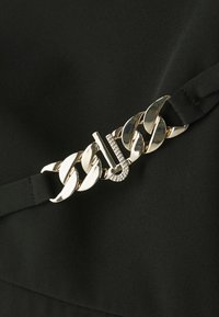 Svart stoff med en metallisk gullkjedeaccent og et diamantrutet logo. Glatt tekstur, minimalistisk design og kontrasterende maskinvare.