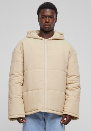 Beige polstret jakke med hette, med glidelås foran og overdimensjonert passform. Stoffet har en glatt tekstur med horisontale sømmer.