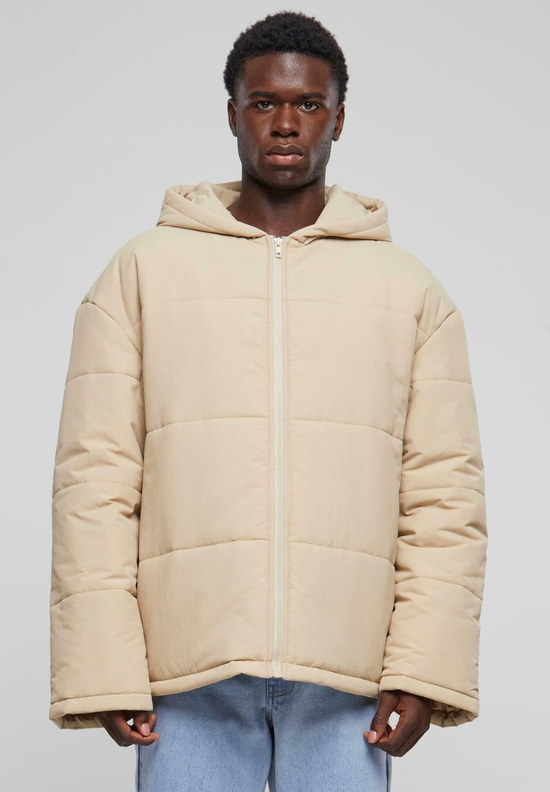 Beige vadderad jacka med huva, med en framsida med dragkedja och oversized passform. Tyget har en slät yta med horisontell sömnad.