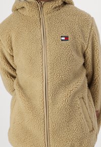 Béžová fleecová bunda se zapínáním na zip vpředu, kapucí a nášivkovým logem. Má strukturovanou látku, dvě boční kapsy a ladící zip.