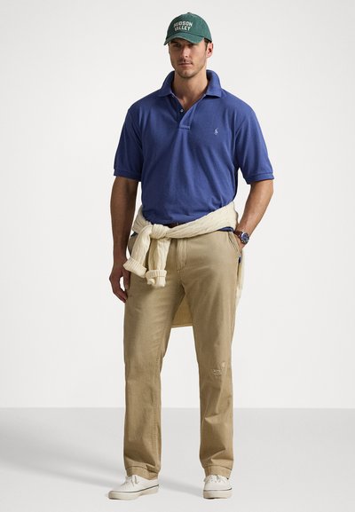 Πόλο μπλουζάκι σε βασιλικό μπλε, κρεμ ζακέτα δεμένη στη μέση, ανοιχτό μπεζ παντελόνι, λευκά αθλητικά παπούτσια, πράσινο καπέλο με κείμενο "Hudson Valley".