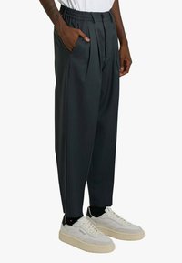 Persona che indossa pantaloni neri plissettati, sneakers bianche con suole beige e calzini neri, in piedi con una mano in tasca.