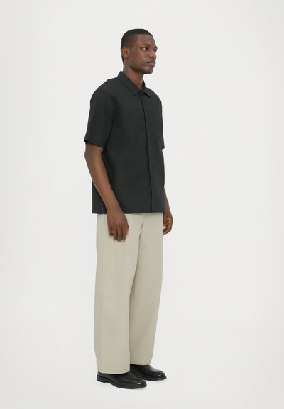 Black short-sleeve button-up shirt, lightweight fabric, square cut. Paired with beige wide-leg trousers and black shoes.