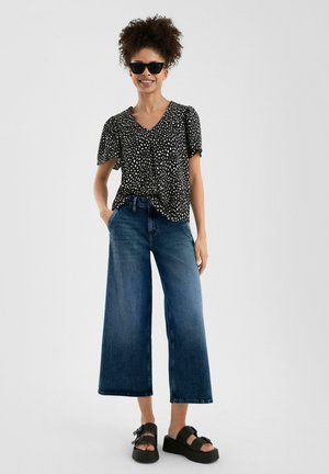 Blouse à pois noir et blanc avec des manches flottantes, associée à un jean bleu en denim à jambes larges et des sandales à plateforme noires.