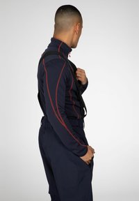 Marineblauw langemouwtop met rode accentstiksel, hoge kraag en een ritssluiting bij de nek; gecombineerd met donkere broeken en een zwarte strap over de schouder.