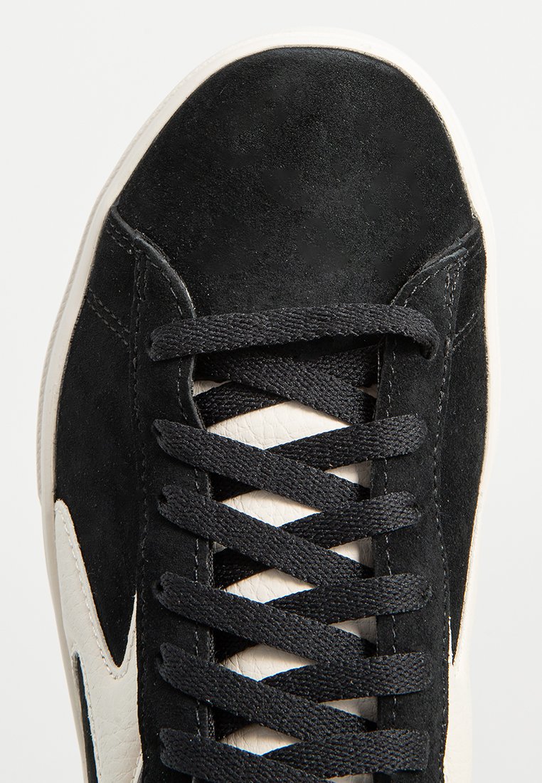 Schwarze Wildledersneaker mit einem beigen Lederlogo, flachen schwarzen Schnürsenkeln und einer strukturierten Oberfläche. Weiße Gummisohle am Boden sichtbar.