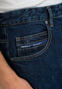 Jeans baggy de mezclilla azul oscuro con un bolsillo trasero que presenta una etiqueta navy distintiva que dice "REELL® BAGGY" y está acentuada con costuras amarillas.