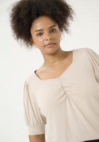 Beige topp med puffärmar, fyrkantig halsringning och samlad detalj i mitten. Mjuk tyg med en slät textur.