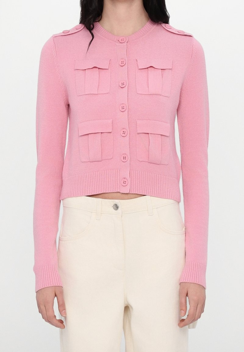 Femme portant un cardigan rose boutonné avec quatre poches avant et un pantalon beige taille haute, sur fond blanc uni.
