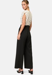 Black wide-leg jumpsuit with a white polka-dot top. The fabric is smooth, with short puffed sleeves and a clean back design. Black heeled sandals.