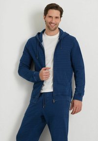 Felpa zipata blu con coulisse regolabile, tasche laterali e una texture morbida, abbinata a una maglietta bianca e pantaloni tuta coordinati.