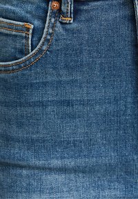 Närbild av blå denimtyg som visar den övre kanten av en frampocket med orange söm och en metallnitar.