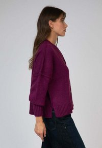 Femme en cardigan en tricot violet et jeans sombres se tient de profil contre un fond gris uni.