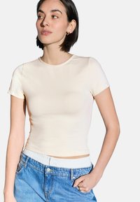 Šviesiaibežinė, prigludusi t-shirt su trumpomis rankovėmis, apvalia iškirpte ir lygia, elastinga medžiaga. Suderinta su aukštu juosmeniu mėlynomis džinsinėmis kelnėmis.