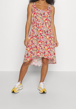 Kvinde iført ærmeløs knælængde kjole med blomsterprint i pink, orange og grønne mønstre, kombineret med gule og hvide sneakers.