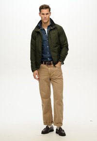 Chaqueta impermeable verde oliva con un alto cuello, llevada sobre una camisa de mezclilla azul. Chinos beige y mocasines oscuros completan el conjunto.