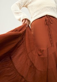 Ruda sluoksniuota maksi sijonas su lengva tekstūra, derinamas su nėriniuotu kreminės spalvos viršumi. Sijonas turi raištelio diržą liemeniai ir švelnias raukšles.