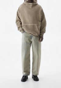 Sudadera beige con capucha y un bolsillo frontal, combinada con vaqueros de mezclilla azul claro de corte holgado. Unos zapatos negros gruesos completan el look.