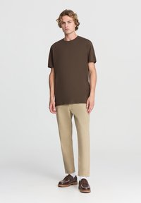 T-shirt corto marrone a maniche corte in cotone, abbinato a pantaloni beige chiaro e scarpe marroni con lacci, esposto su una figura maschile in piedi.