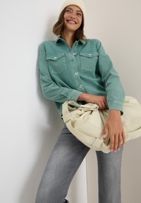 Femme souriante portant un bonnet crème, une chemise bleu canard boutonnée et un jean gris, tenant un grand sac beige, s'appuyant contre un mur bicolore.