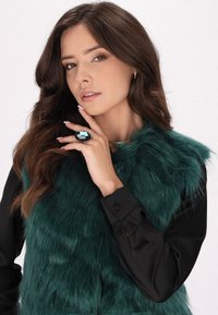 Grüne Kunstpelz-Weste mit schwarzem Langarmbluse. Das Model trägt einen großen ovalen Ring mit blauen und schwarzen Akzenten. Strukturiertes und geschichtetes Erscheinungsbild.