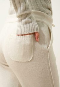 Krämfärgade, mjuka stickade byxor med ribbad midja, utrustade med ett sidofack och matchande manschetter. Slät textur med minimala sömmar.