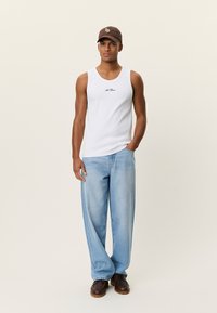 Débardeur blanc avec logo noir, associé à un jean large bleu clair et des chaussures marron. La casquette est marron avec un détail brodé.