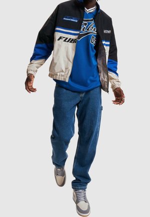 Homme marchant vers l'avant portant une veste de course FUBU bleue et beige, un jean bleu, des baskets grises et blanches et un t-shirt bleu FUBU.