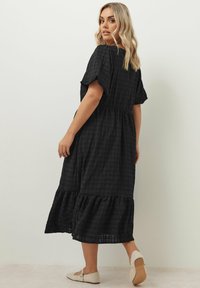 Musta midi-mekko, joka on valmistettu teksturoidusta kankaasta, lyhyillä hihoilla, vyötärön kireydellä ja levenevällä helmalla, jossa on röyhelöyksityiskohtia.