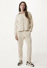Beige rits-jack met een kraag, gecombineerd met bijpassende tapse broek. Gestructureerde stof, ronde zakken en sneakers van gemengd materiaal met accenten.