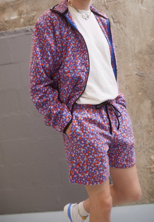 Person trägt eine passende lila, rote und blaue gemusterte Jacke und Shorts mit weißem T-Shirt und weißen Sneakers mit blauen Akzenten.