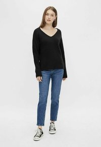 Young woman standing, wearing a black V-neck sweater, blue jeans, and black-and-white sneakers against a plain light background.