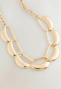 Fette Goldkette mit abwechselnd glatten, länglichen ovalen und halbmondförmigen Gliedern auf einem hellen Hintergrund.