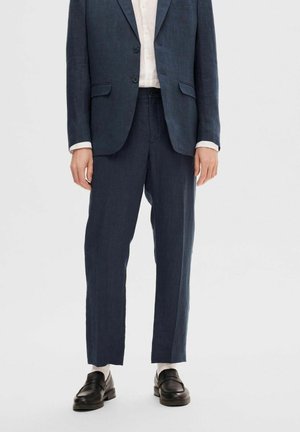 Completo in lino blu scuro con texture, dotato di blazer su misura e pantaloni a gamba dritta. Abbinato a una camicia chiara e mocassini neri.