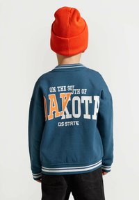 Niebieska bluza z napisami "NA POŁUDNIU DAKOTY, STAN USA" w pomarańczowym i białym kolorze, ściągacz przy kołnierzu i mankietach, oraz jaskrawopomarańczowa czapka.