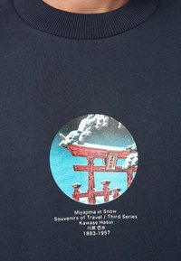 Tamsiai mėlynas džemperis su apvaliu raštu, kuriame pavaizduotas raudonas torii vartai sniege, su tekstu „Miyajima sniege“ ir menininko vardu po juo.