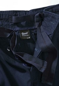 Granatowe spodnie z teksturowaną tkaniną, regulowanym paskiem w pasie, dwoma guzikami oraz metką oznaczającą rozmiar M i markę "Brandit".